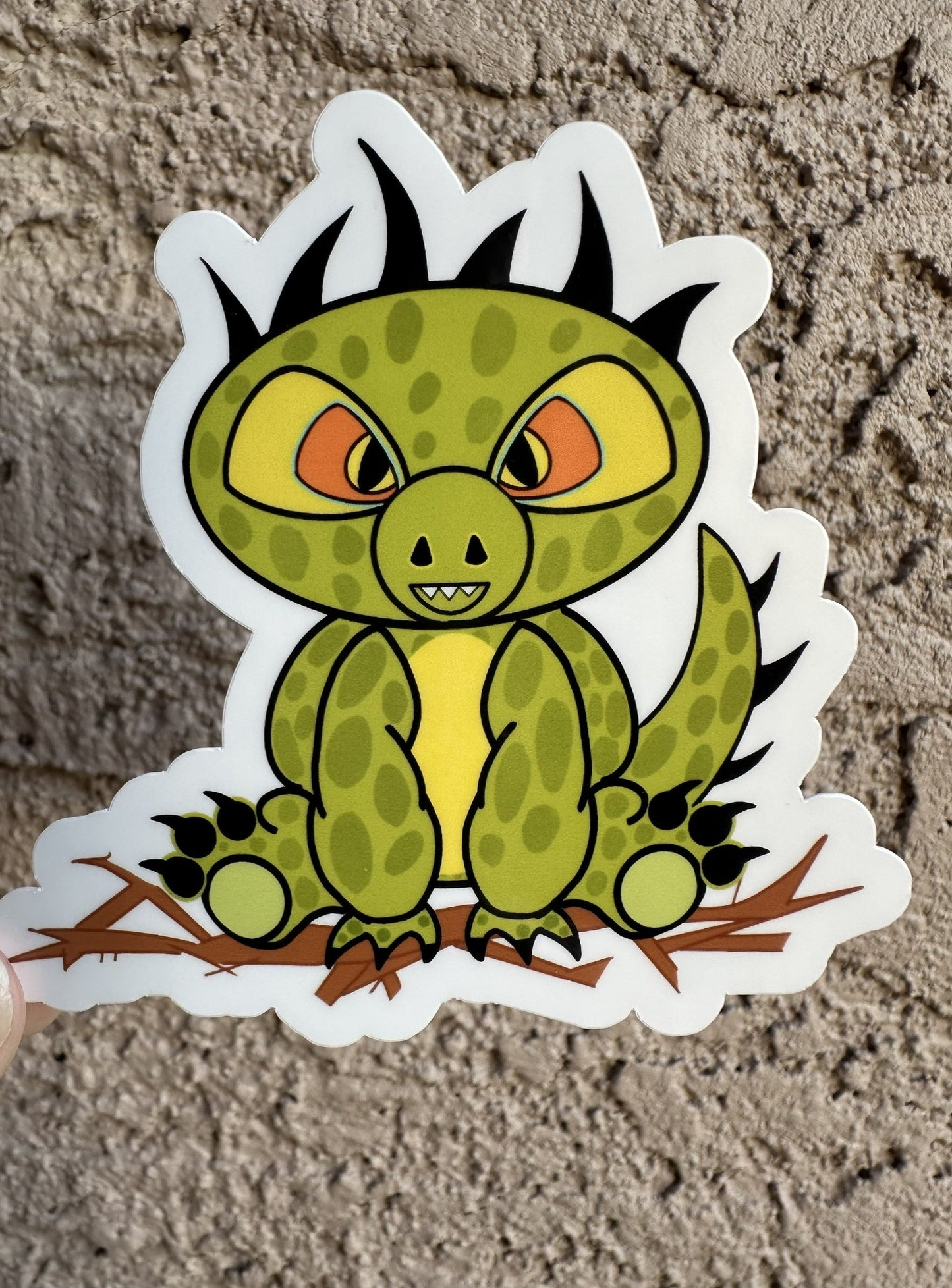Cartoon half dinosaur half lizard sticker. The sticker character is green and light lime in the middle of its stomach. The character also has dark green dots with black hair and has black claws coming from the hands and feet. The character has orange and black eyes. The character is sitting on top of brown branches. The sticker background is on a textured wall.