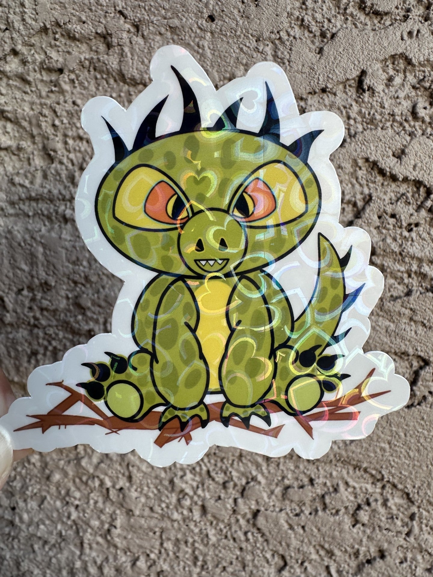 Heart holographic cartoon half dinosaur half lizard sticker.  sticker character is green and light lime in the middle of its stomach. The character also has dark green dots with black hair and has black claws coming from the hands and feet. The character has orange and black eyes. The character is sitting on top of brown branches. The sticker background is on a textured wall.