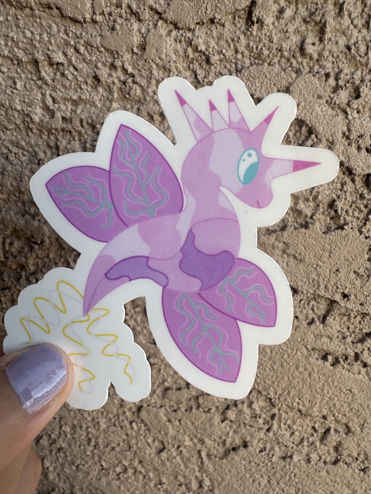 Fictional character sticker that is light purple and dark purple, that looks like a dragon and also has wings with turquoise lines. The character has five horns coming from its head that are pink and purple, while the tail is yellow and light pink. The character has light blue eyes. 