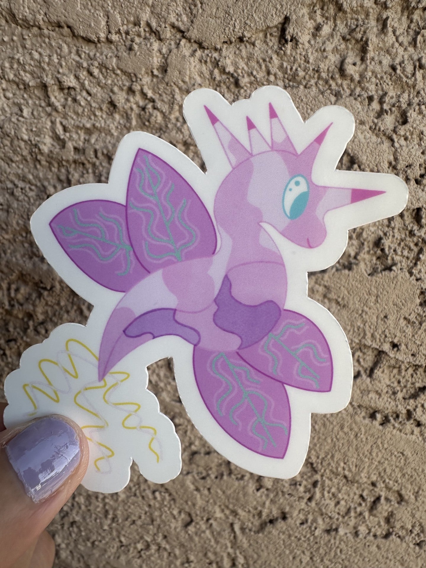 Fictional character sticker that is light purple and dark purple, that looks like a dragon and also has wings with turquoise lines. The character has five horns coming from its head that are pink and purple, while the tail is yellow and light pink. The character has light blue eyes. 