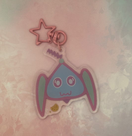 Fictional character keychain that is light blue and turquoise that has big and white eyes, with a purple antenna on head and has a cookie next to it. The keychain has a pink star key ring. The acrylic keychain is sitting on top of a pink textured background.