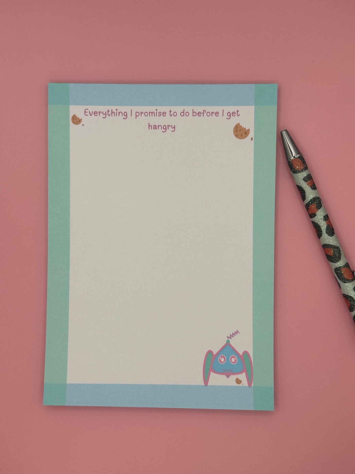 A notepad that has fictional character at the bottom that is blue, pink and turquoise and has an antenna coming from it. The character is next to a cookie. The notepad edges all around has a border color of turquoise and blue and has a heading on top with cookies stating, "Everything I promise to do before I get hangry". The notepad is sitting on top of a pink background with a pen. 
