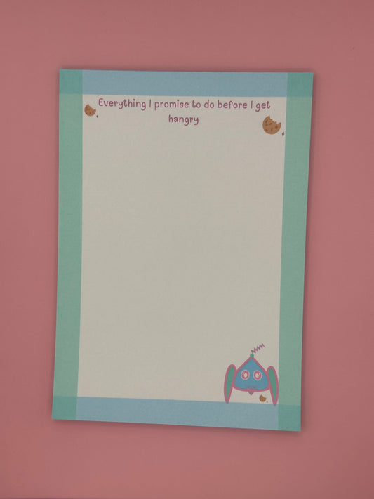 A notepad that has fictional character at the bottom that is blue, pink and turquoise and has an antenna coming from it. The character is next to a cookie. The notepad edges all around has a border color of turquoise and blue and has a heading on top with cookies stating, "Everything I promise to do before I get hangry". The notepad is sitting on top of a pink background with a pen. 
