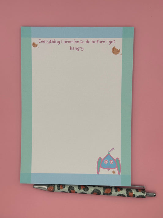 A notepad that has fictional character at the bottom that is blue, pink and turquoise and has an antenna coming from it. The character is next to a cookie. The notepad edges all around has a border color of turquoise and blue and has a heading on top with cookies stating, "Everything I promise to do before I get hangry". The notepad is sitting on top of a pink background with a pen. 