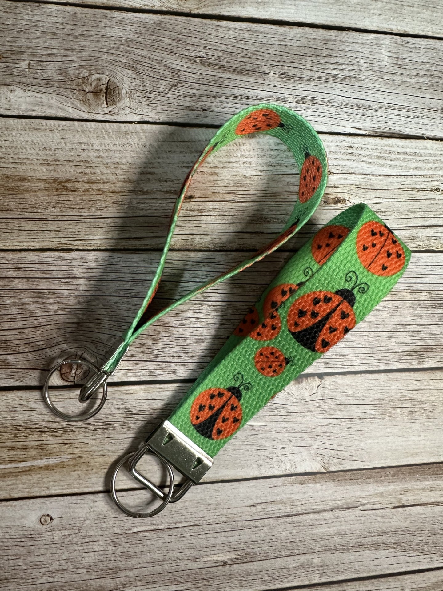 A fabric wristlet keychain featuring a green background patterned with black and red ladybugs, shown on a light wooden surface.