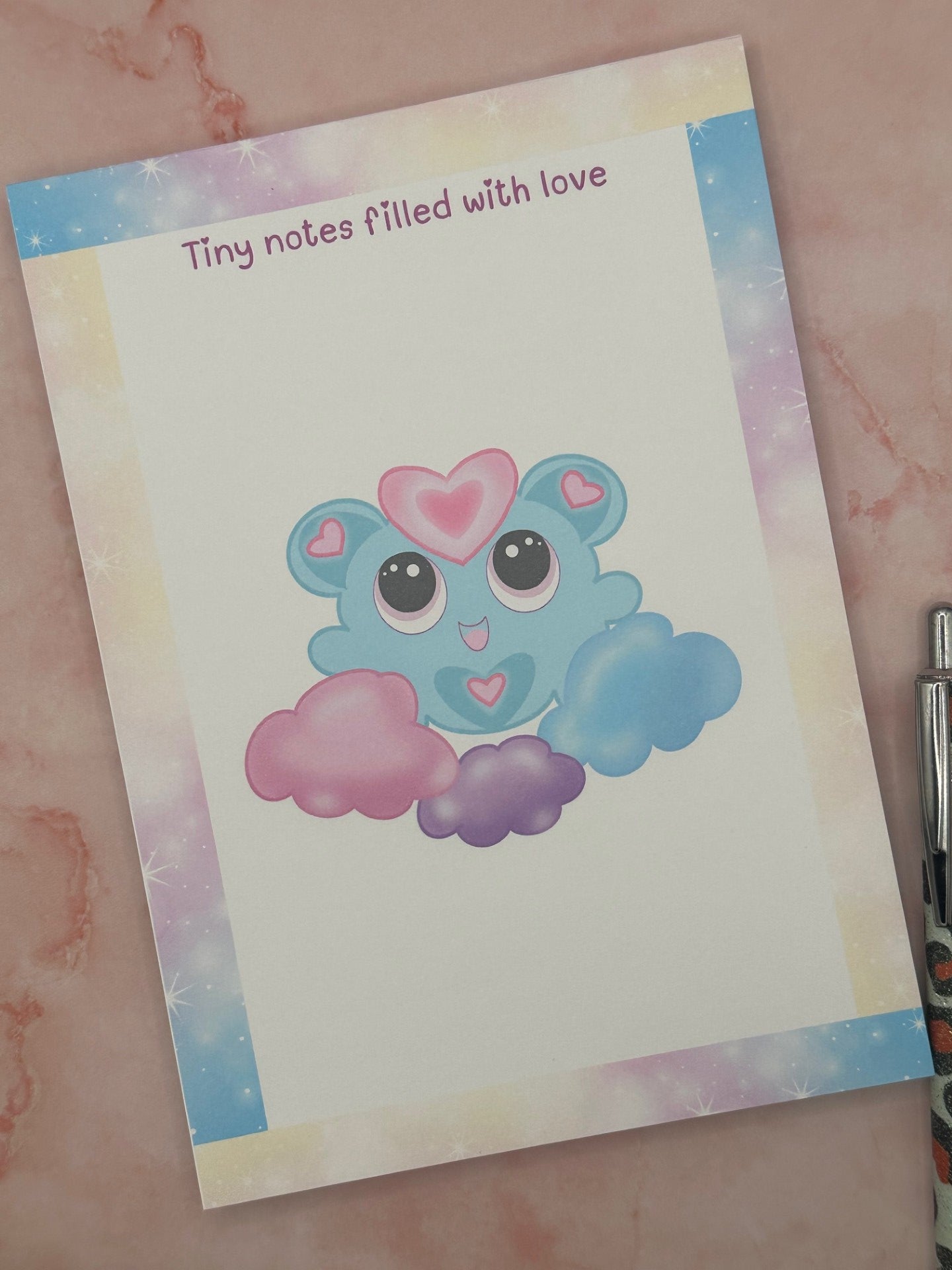 Notepad that has pastel sparkle colors all around the four edges. The notepad heading states "Tiny notes filled with love". The character has a fictional character on it that is blue and pink hearts on the ears and stomach. The character is floating on blue, purple, and pink clouds. The notepad is sitting on top of a pink background. 