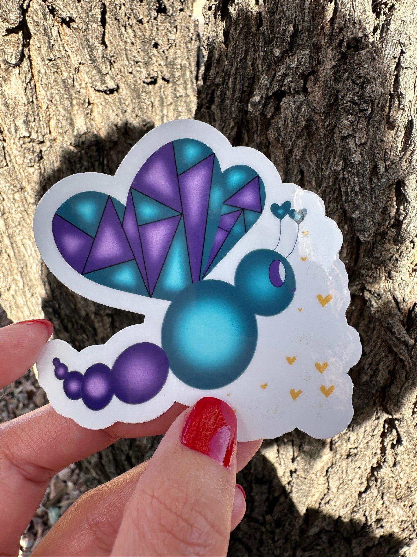 Colorful fictional character butterfly-shaped sticker held by a hand with red nail polish against a tree bark background