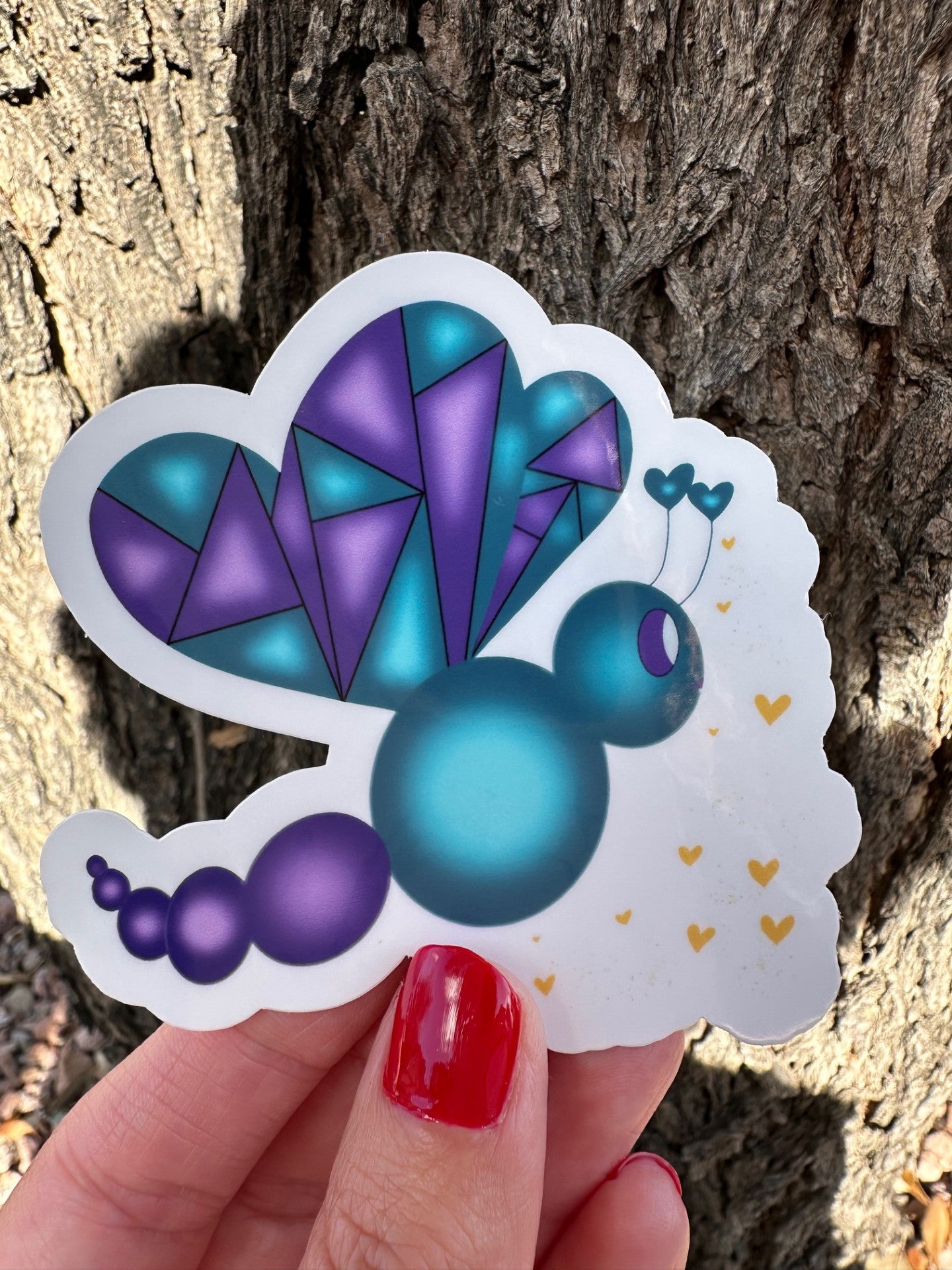 Colorful fictional character butterfly-shaped sticker held by a hand with red nail polish against a tree bark background