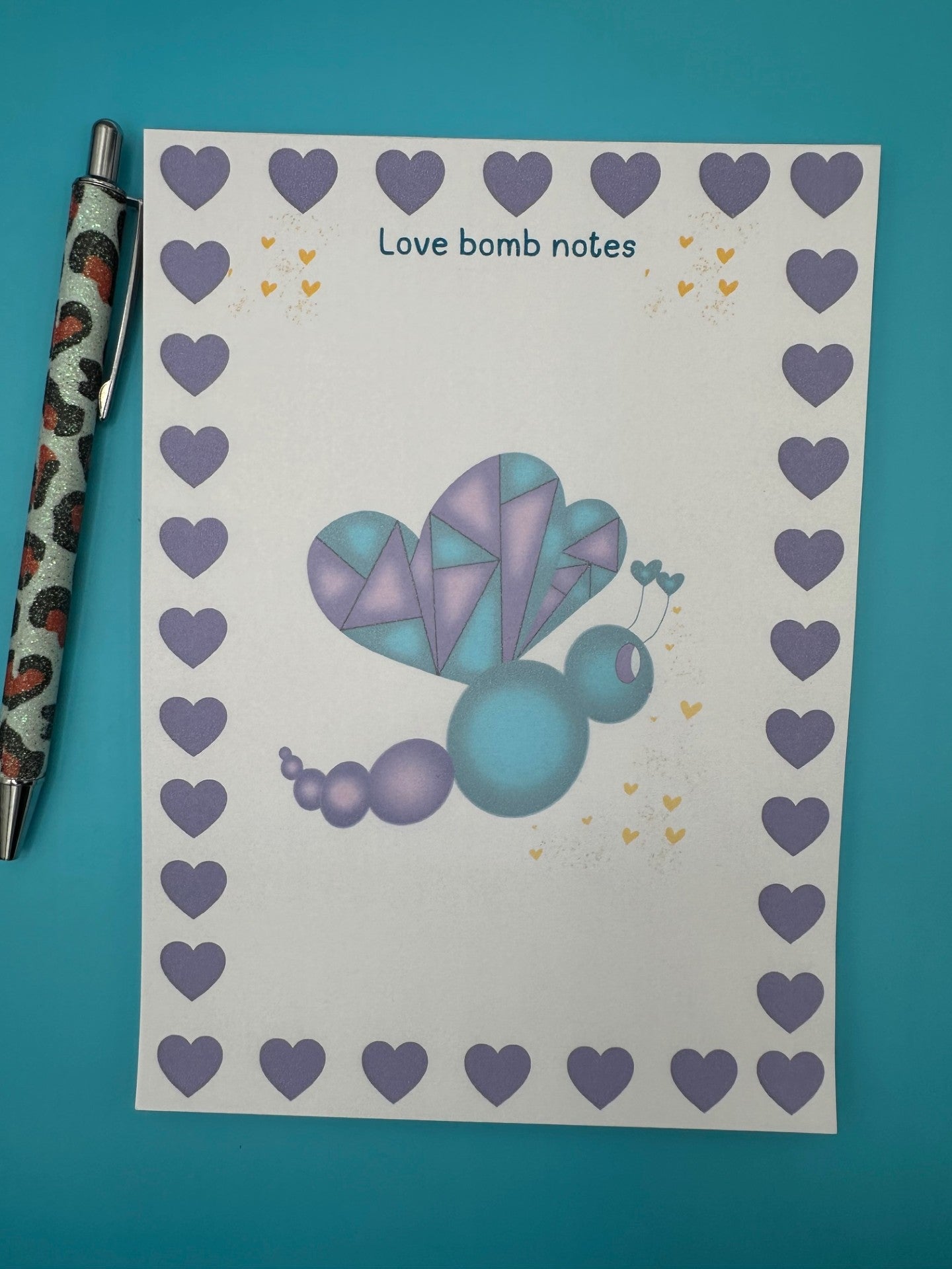 A notepad with a colorful butterfly illustration with purple and dark blue colors and a heart border. The notepad is on a blue background, accompanied by a pen.