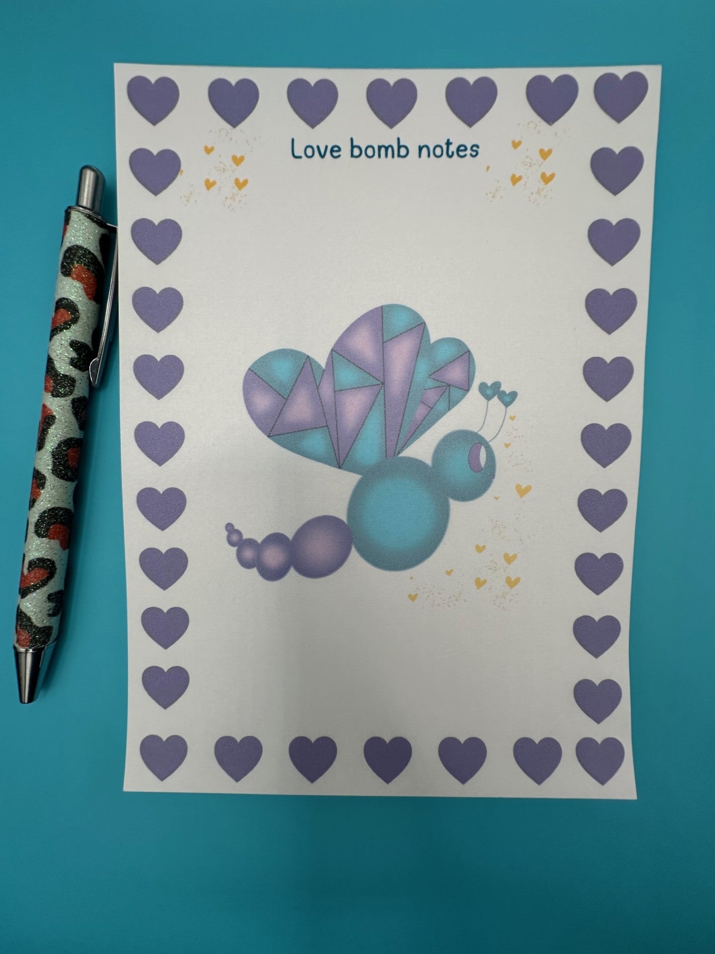 A notepad with a colorful butterfly illustration with purple and dark blue colors and a heart border. The notepad is on a blue background, accompanied by a pen.