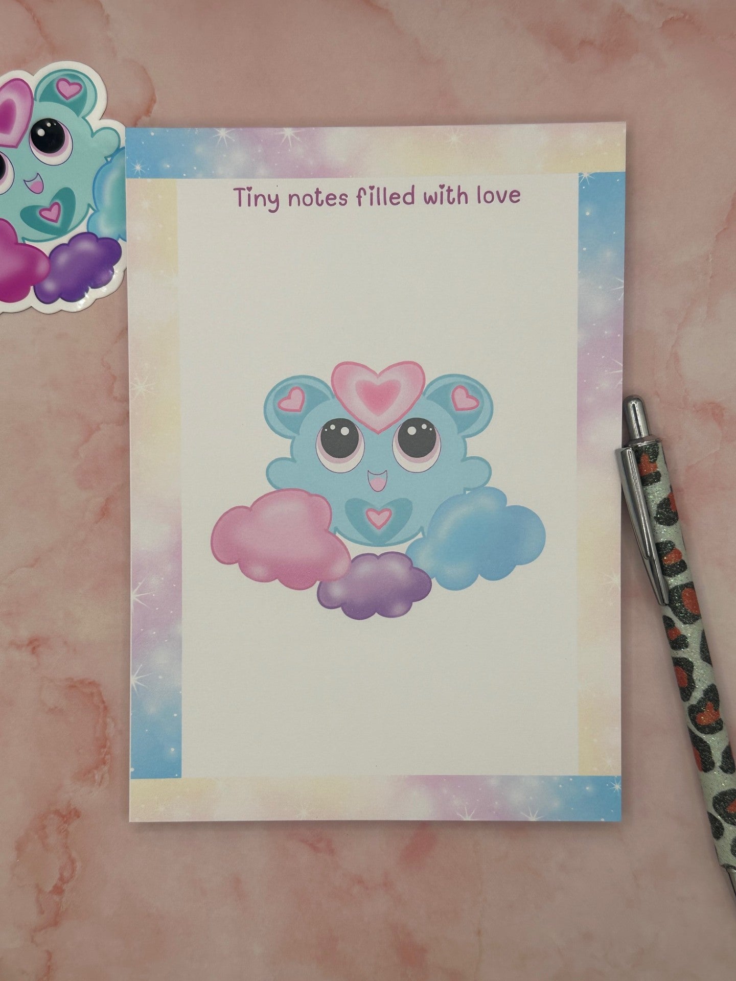 Notepad that has pastel sparkle colors all around the four edges. The notepad heading states "Tiny notes filled with love". The character has a fictional character on it that is blue and pink hearts on the ears and stomach. The character is floating on blue, purple, and pink clouds. The notepad is sitting on top of a pink background with a pen and a sticker.