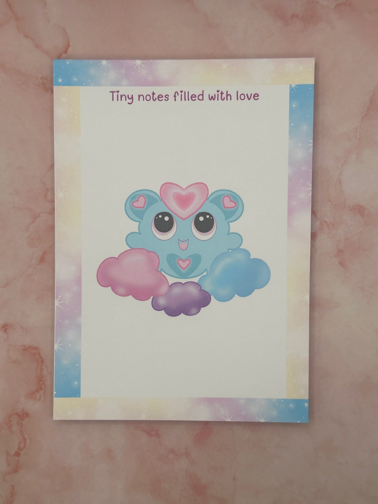 Notepad that has pastel sparkle colors all around the four edges. The notepad heading states "Tiny notes filled with love". The character has a fictional character on it that is blue and pink hearts on the ears and stomach. The character is floating on blue, purple, and pink clouds. The notepad is sitting on top of a pink background. 