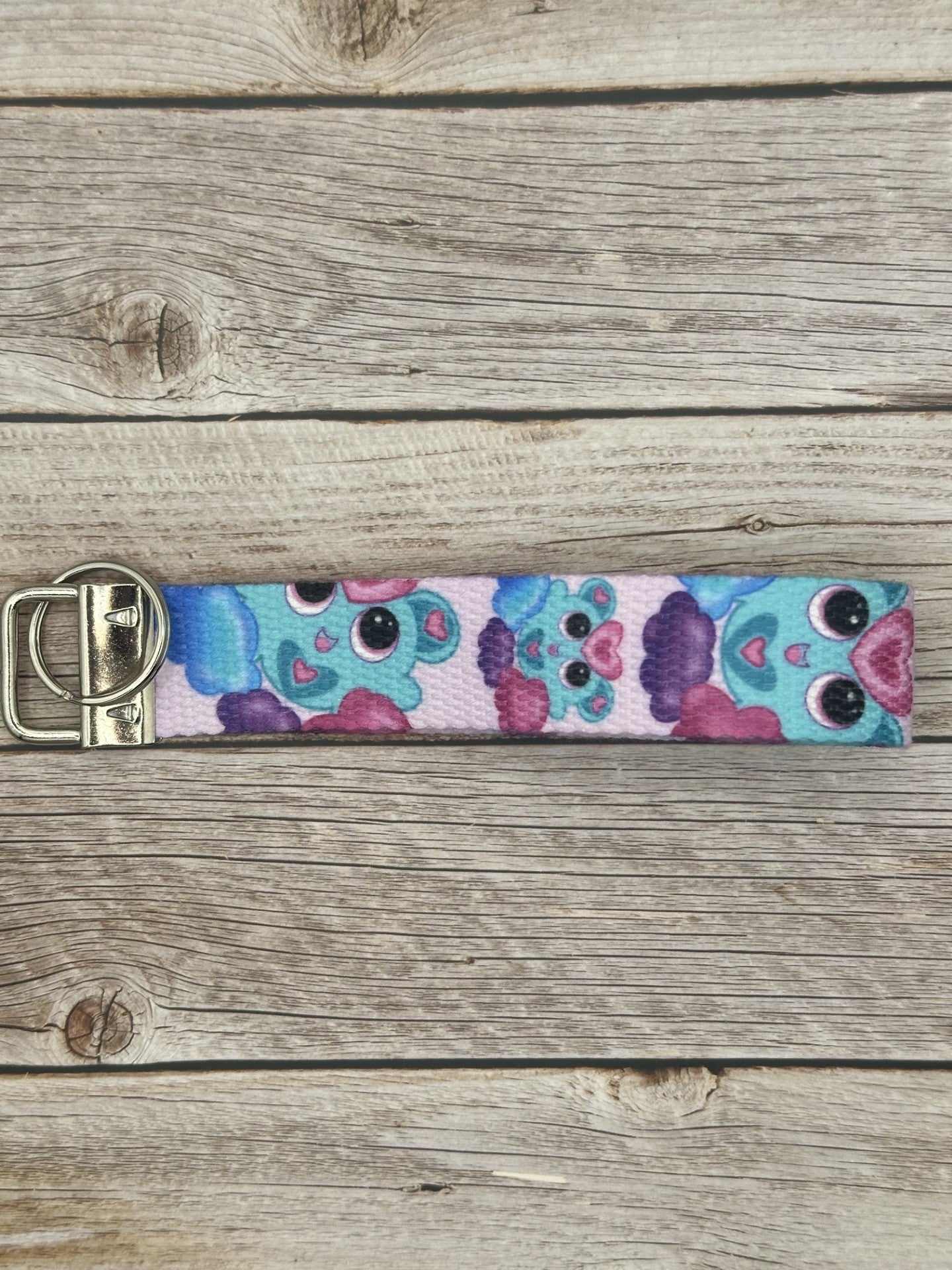 Unique fictional character that is blue and has pink and turquoise hearts on it. The character is also on a purple, pink and blue cloud and has black eyes with pink in them. The background color of the wristlet is light pink. The wristlet is sitting on top of a wooden background. 