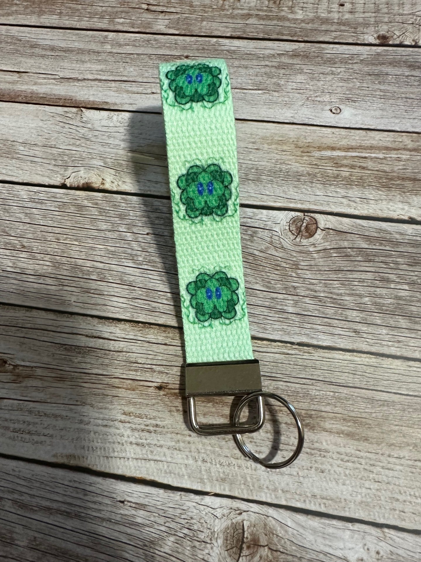 Green keychain strap with floral patterns on a wooden surface, with a fictional character, Lucky, with his vines up.