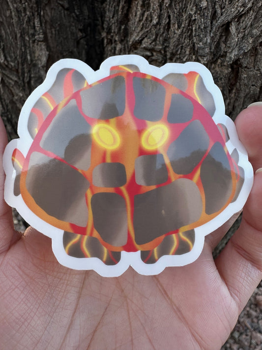 Sticker of a unique creature with fiery lava design and bright yellow eyes held in a hand against a tree bark background.
