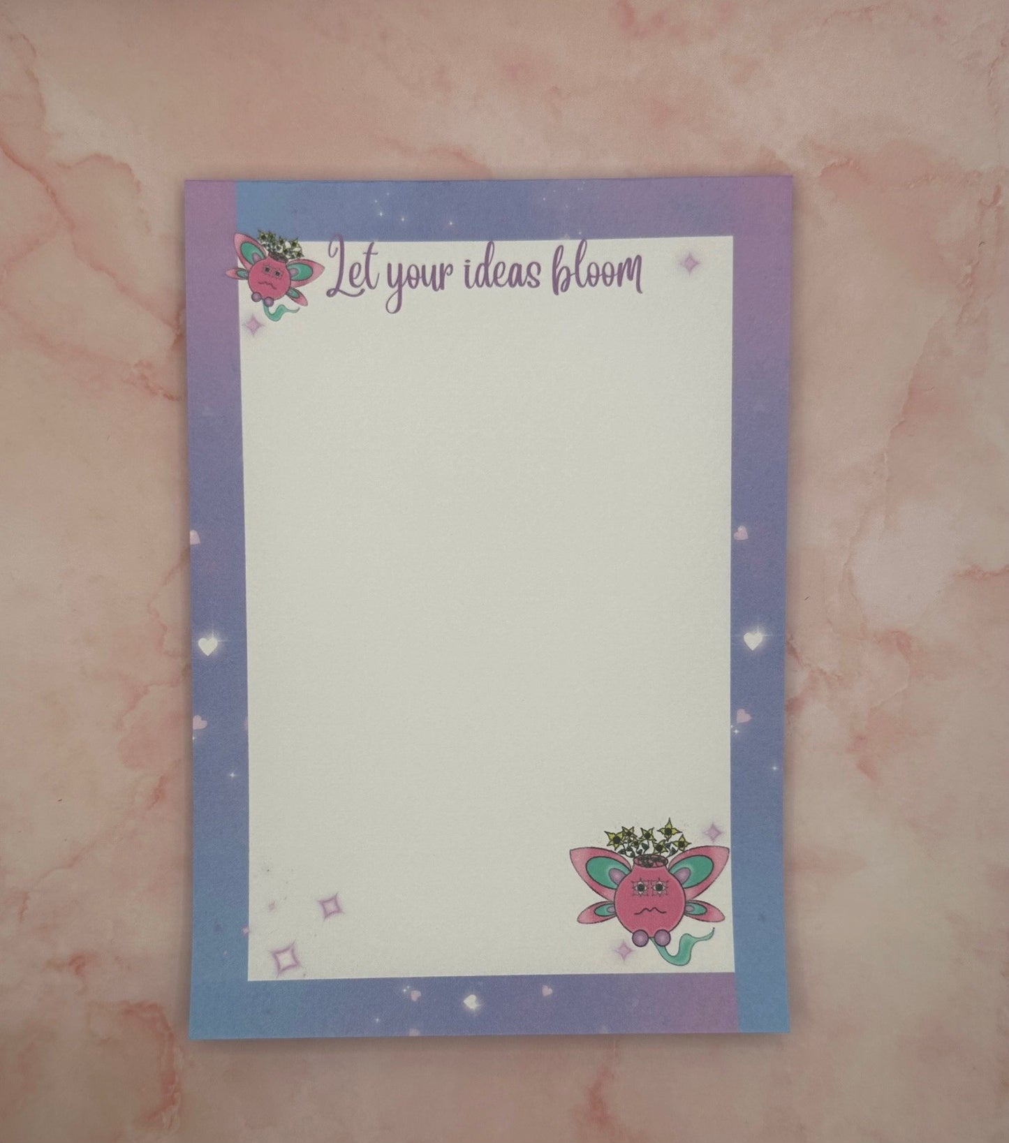 A notepad with fictional characters on top and bottom that are pink and turquoise color having yellow flowers come out of it. Around the notepad is purple color with hearts. The notepad heading states "let your ideas bloom". While the notepad is sitting on a pink background with a pen. 