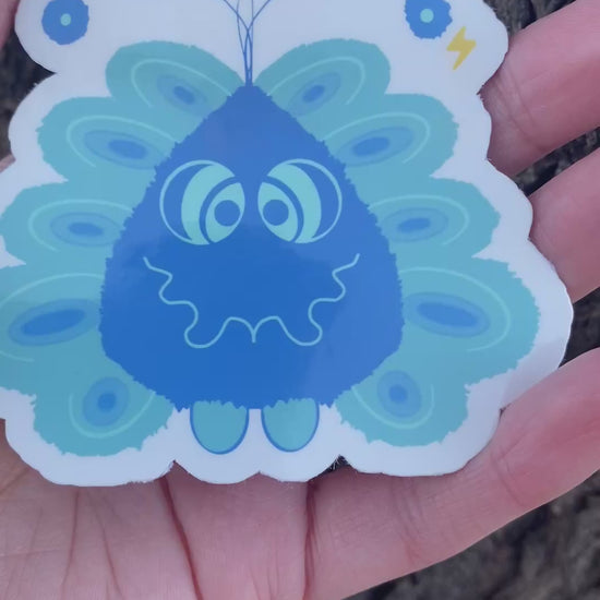 A sticker featuring a friendly blue cartoon monster character with a wavy smile, antenna with spheres, peacock-like feather details, and small yellow lightning bolts.