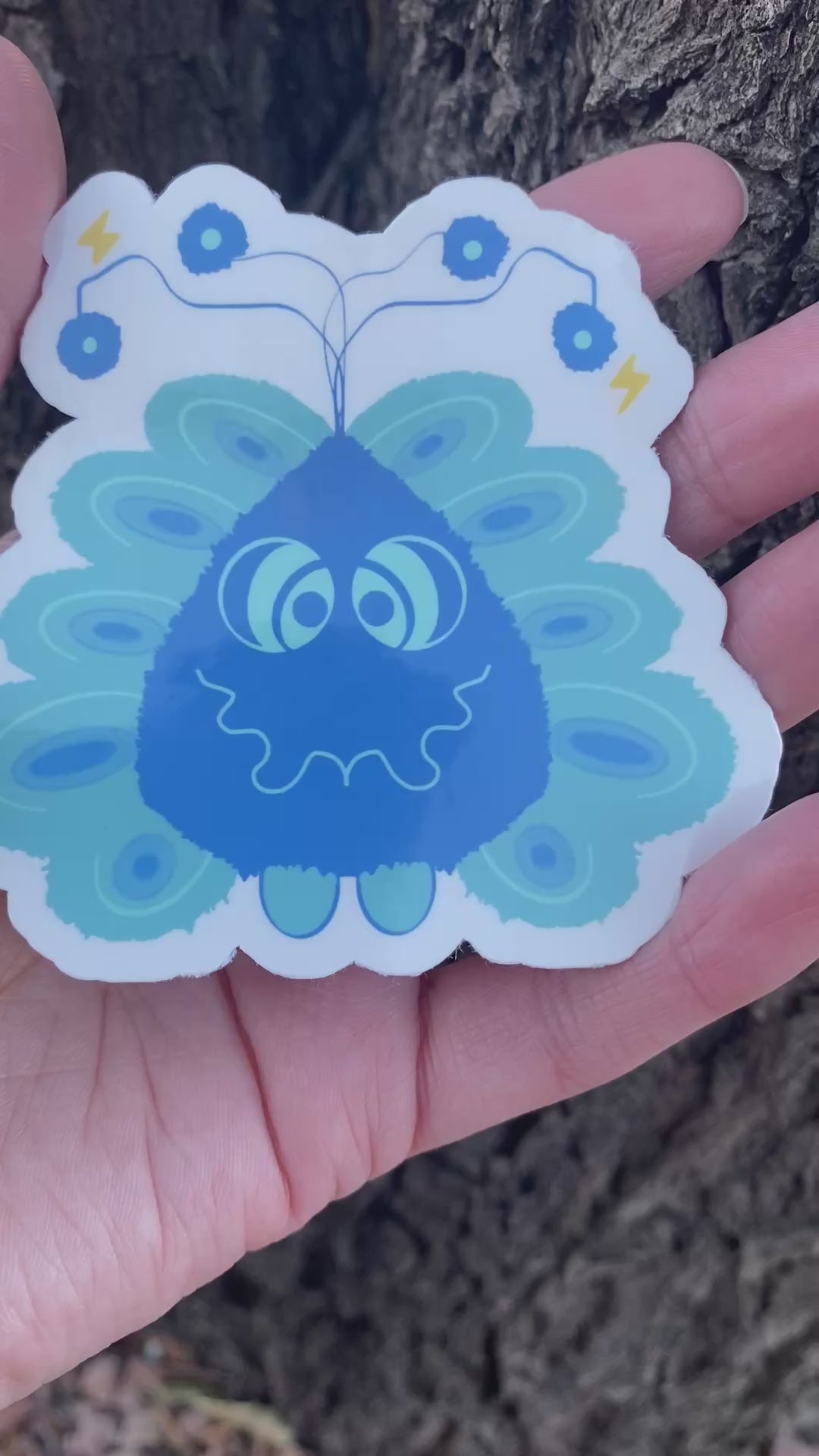 A sticker featuring a friendly blue cartoon monster character with a wavy smile, antenna with spheres, peacock-like feather details, and small yellow lightning bolts.