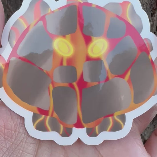 Sticker of a unique creature with fiery lava design and bright yellow eyes held in a hand against a tree bark background.