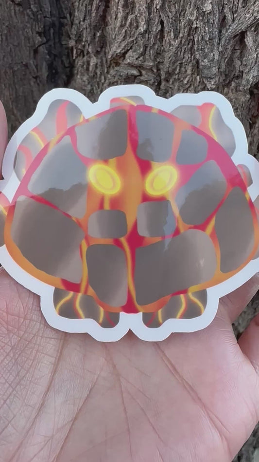 Sticker of a unique creature with fiery lava design and bright yellow eyes held in a hand against a tree bark background.