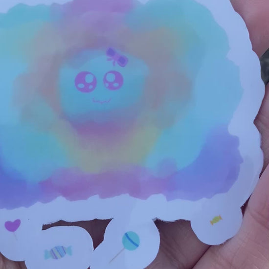A sticker of a colorful, watercolor-style cloud shape in pastel blue, pink, yellow, and purple, featuring a cartoon face in the center and small candy illustrations along the bottom edge.