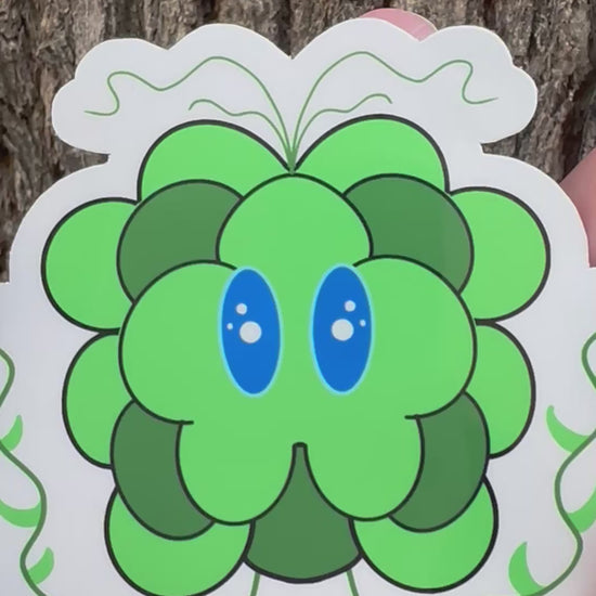 Green cartoonist character with blue eyes and small decorative vine illustrations held against a tree bark background.