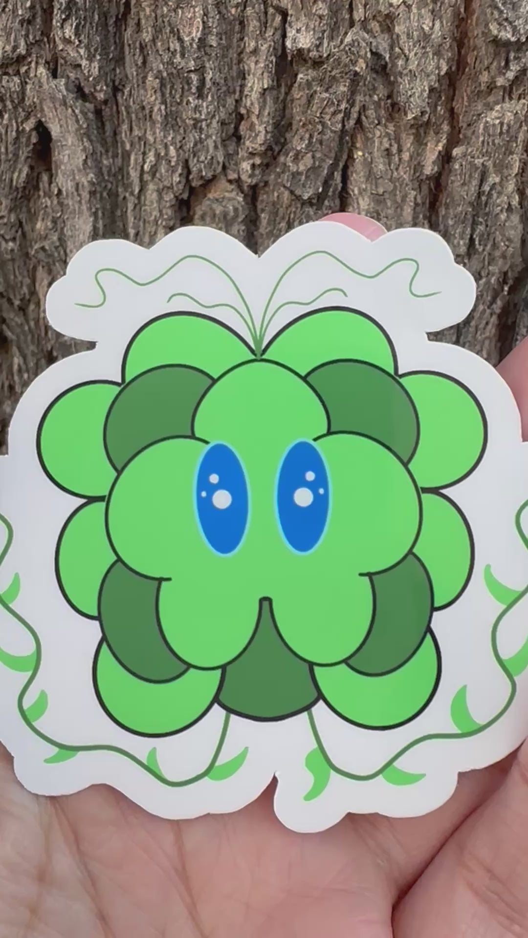 Green cartoonist character with blue eyes and small decorative vine illustrations held against a tree bark background.