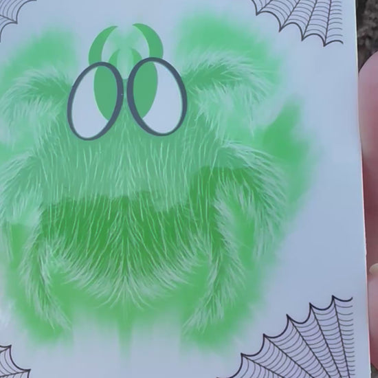 A square sticker featuring a fluffy green cartoon spider with large black and white eyes. Black spider web illustrations decorate all four corners.