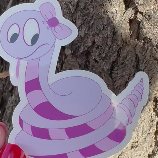 Video of a Cartoon non-fiction character snake shape with a purple color sticker held against a tree bark background.