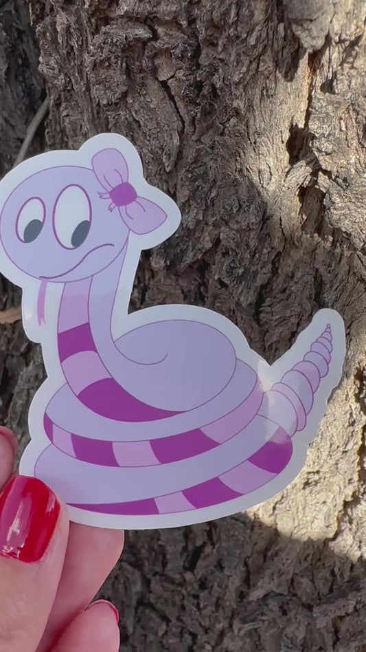 Video of a Cartoon non-fiction character snake shape with a purple color sticker held against a tree bark background.