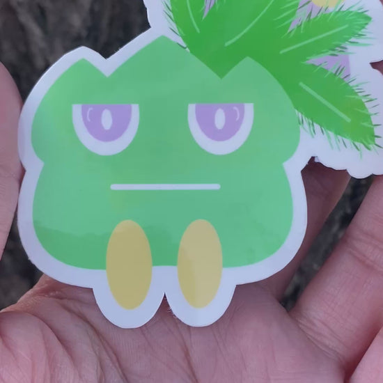 Cartoonish green creature sticker held by a hand against a tree bark background.