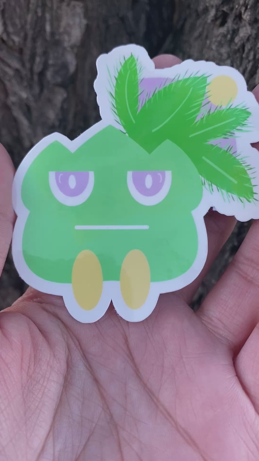 Cartoonish green creature sticker held by a hand against a tree bark background.