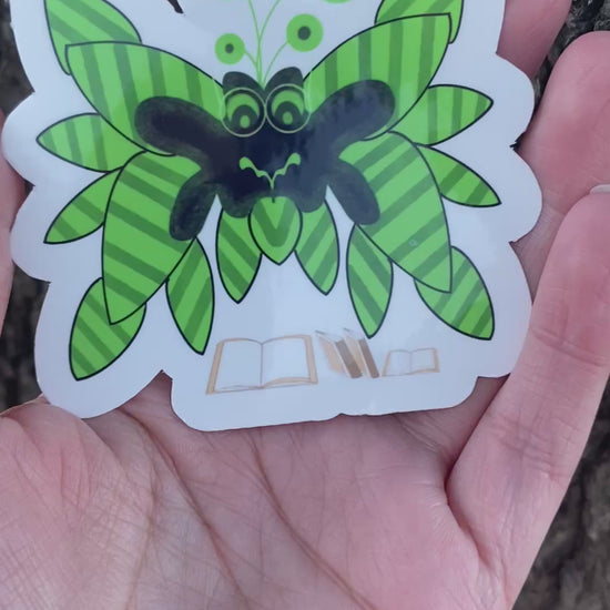 A sticker featuring a cartoon unique creature design with green striped wings and a black body. Below the moth are small illustrations of open and closed books.