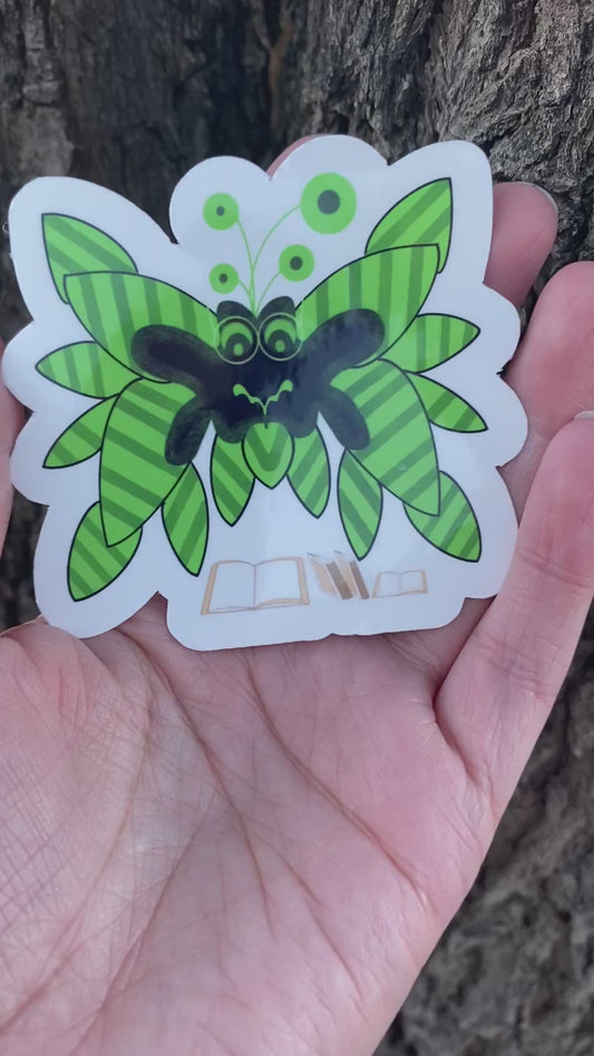 A sticker featuring a cartoon unique creature design with green striped wings and a black body. Below the moth are small illustrations of open and closed books.