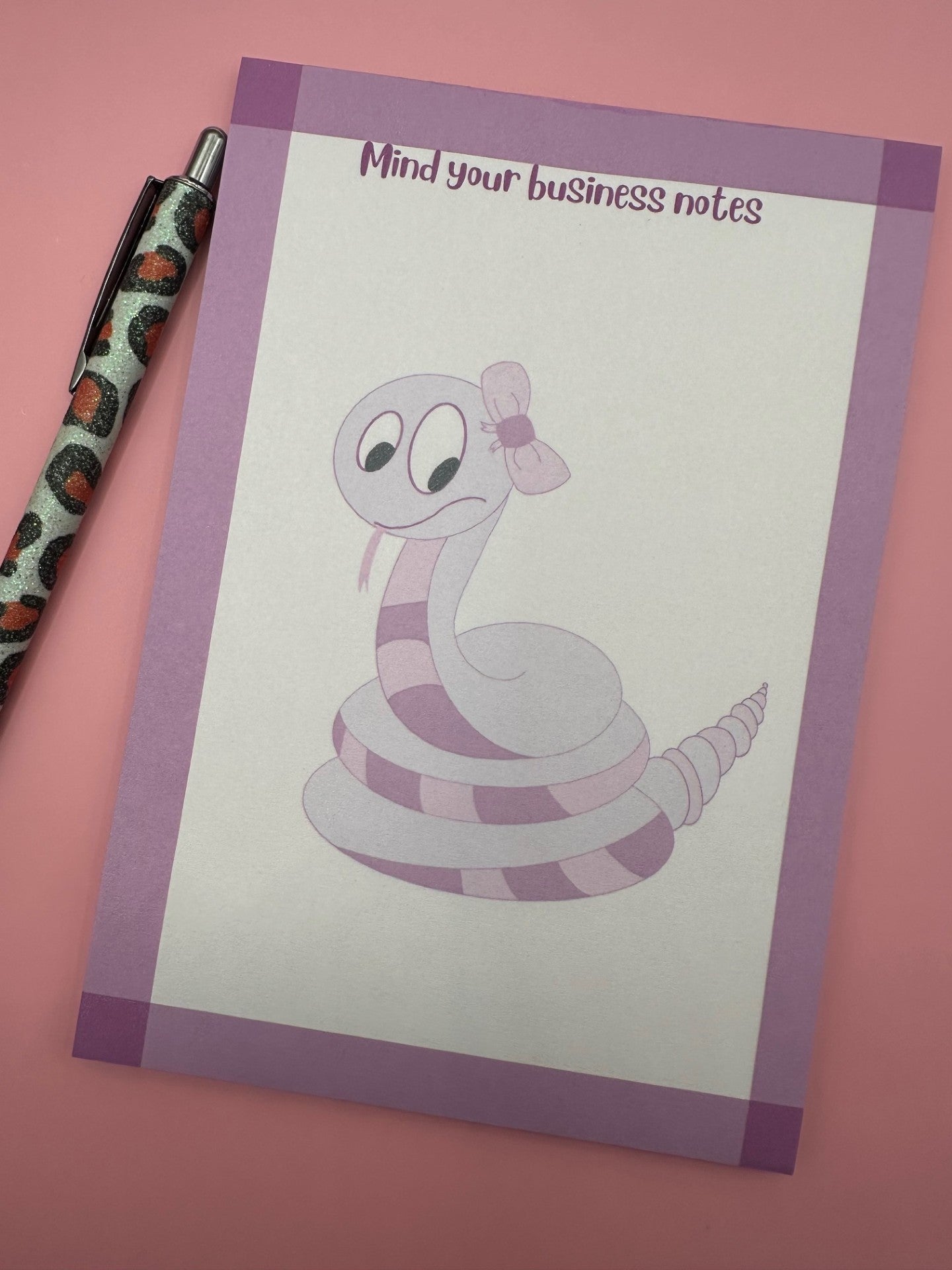 A notepad with fictional character is that a dark and light pink color with a bow on top of the head and the tongue sticking out. The notepad has purple edges and the notepad heading stating "mind your business notes". 