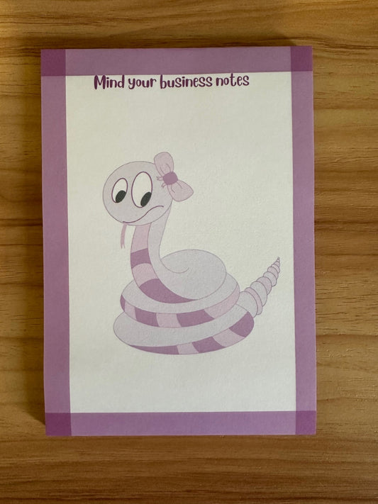 Notepad with a hand-drawn cartoon snake design that is purple, with a heading on the notepad that states "mind your business " on a wooden surface.