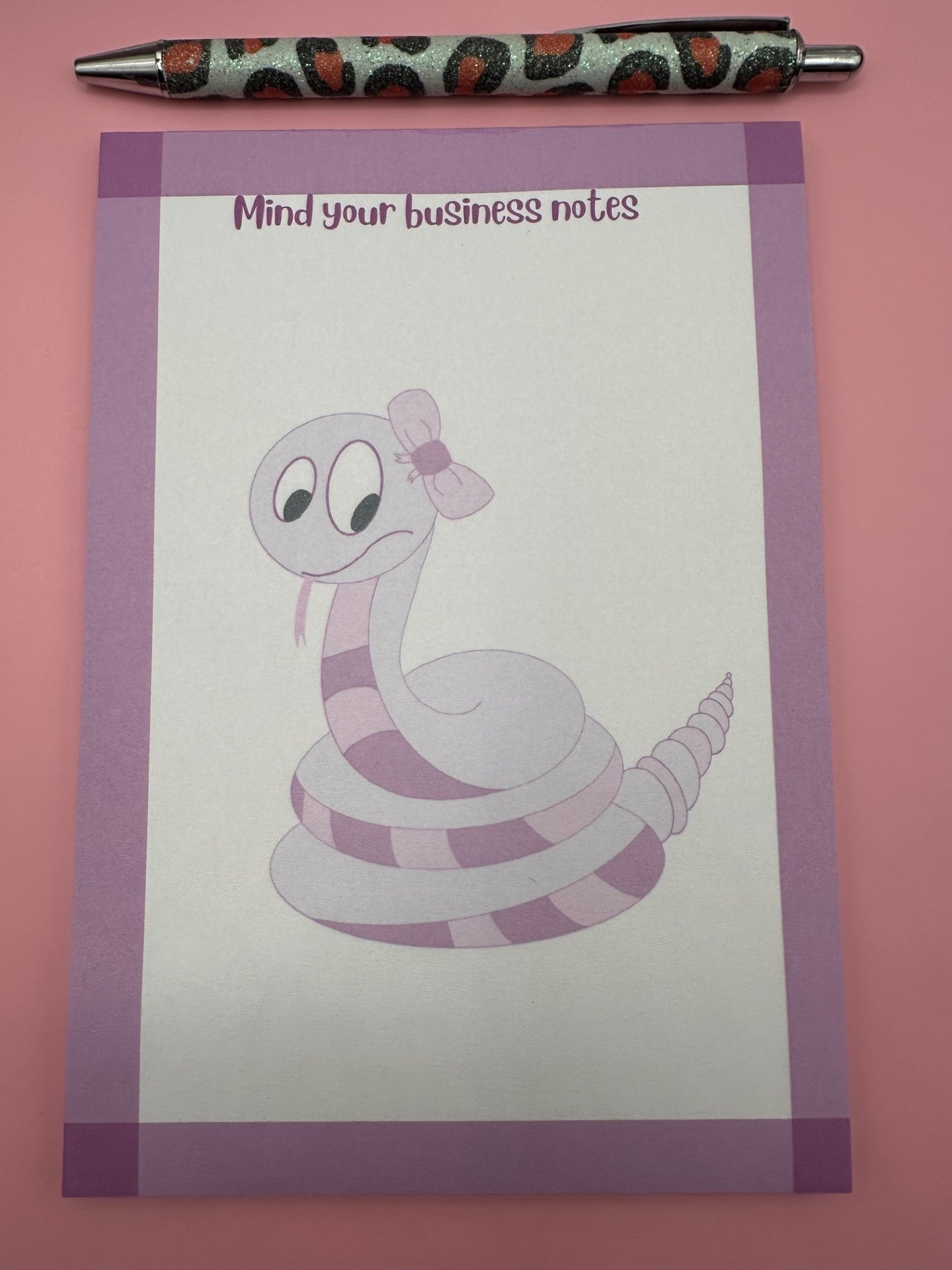 A notepad with fictional character is that a dark and light pink color with a bow on top of the head and the tongue sticking out. The notepad has purple edges and the notepad heading stating "mind your business notes". 