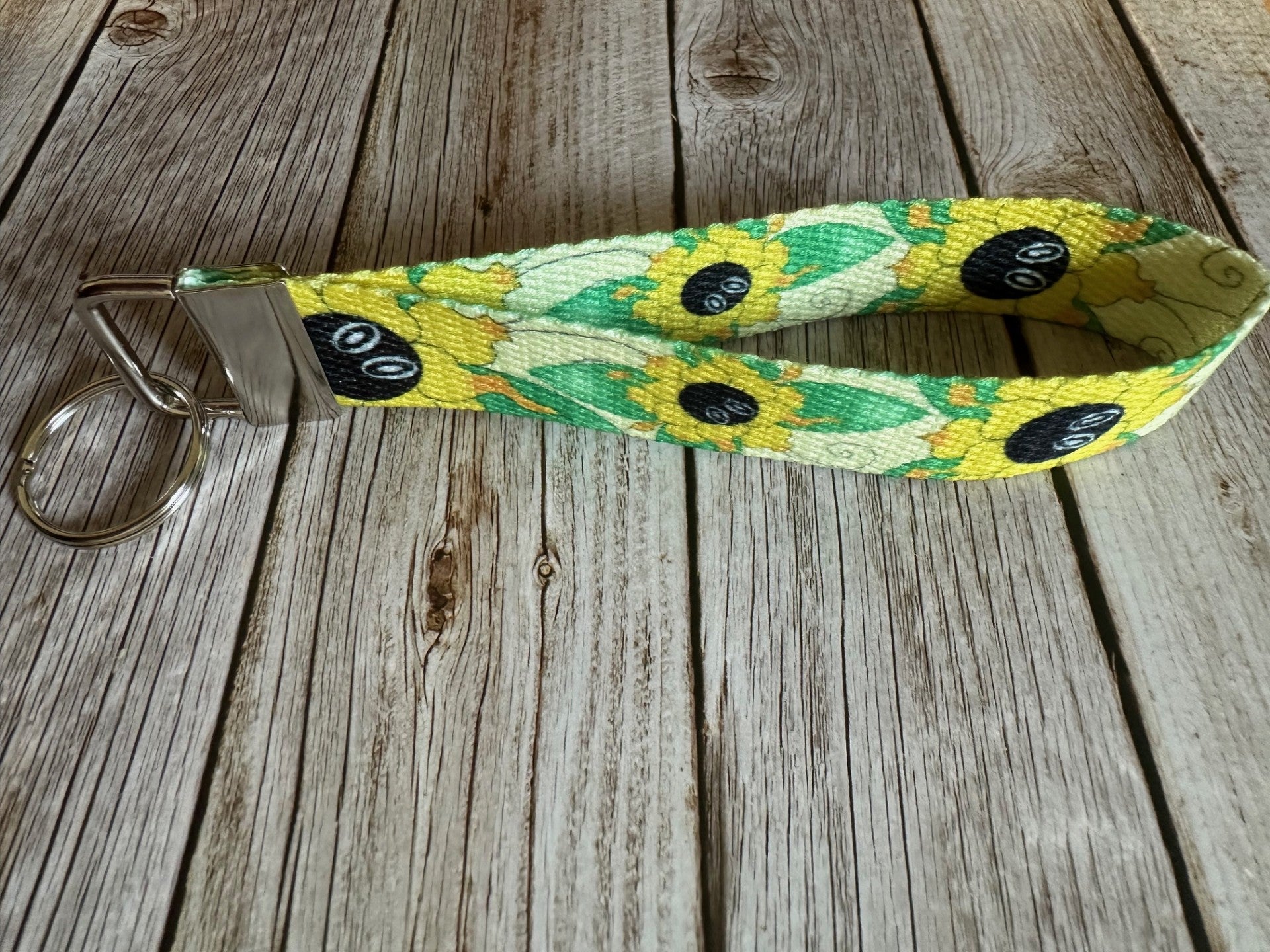 Keychain wristlet with a yellow flower that also has green butterfly wings coming from it. The wristlet is sitting on a wooden surface. 